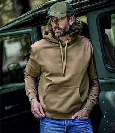 Hoodies Man in a brown hoodie and cap leans against a vehicle in a forested setting.