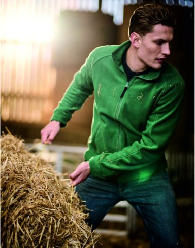 Fleeces Man in a green jacket stacking hay in a barn with warm lighting.